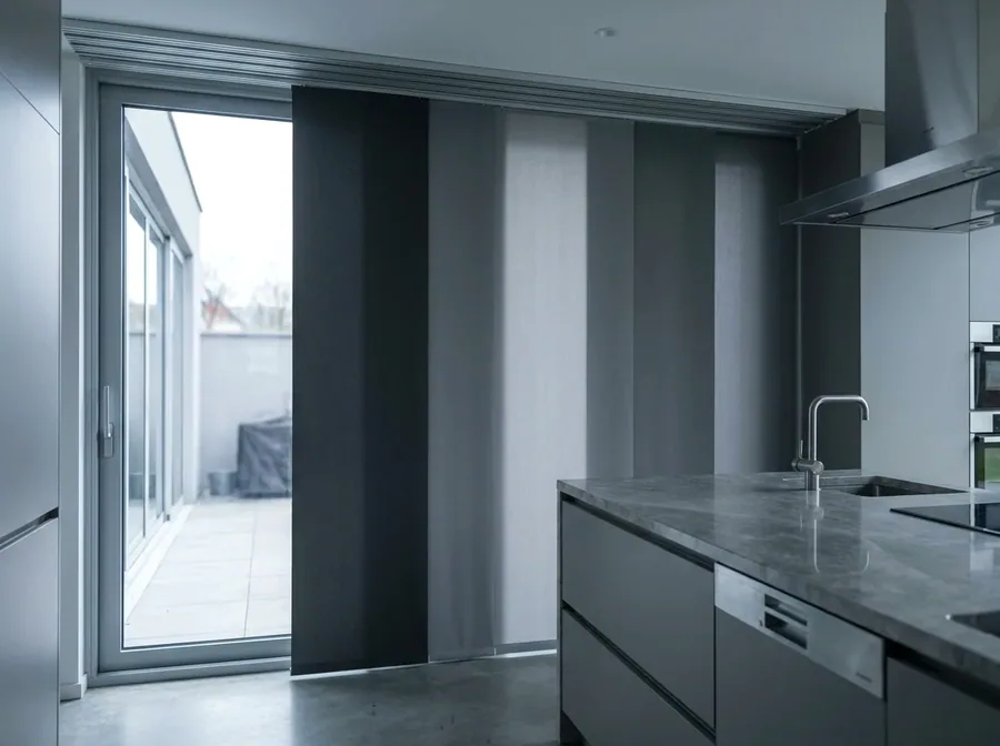 Wide sliding fabric panel blinds tracking across a glazed wall in a modern kitchen